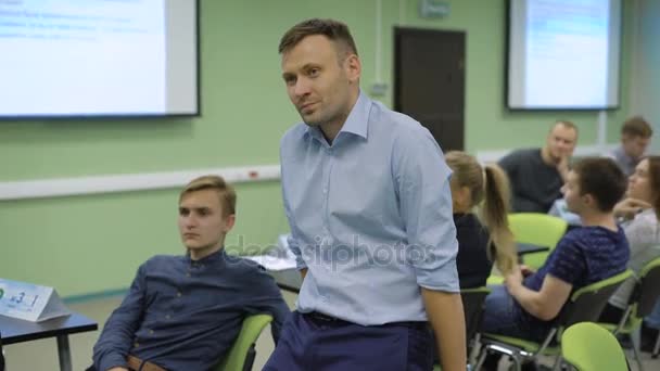 Mladý učitel ve třídě stojí opřen lokty o stůl a mají se studenty. Muž oblečený v modré košili se klást otázky z publika. — Stock video