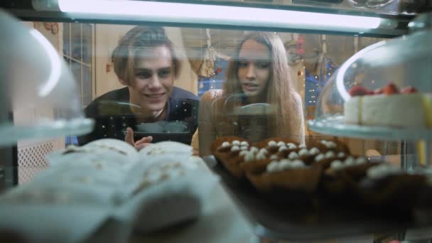 Beau couple de jeunes dans un magasin de bonbons. Une fille et un mec en pull choisissent Gâteau et desserts pour eux-mêmes. Vitrine agréablement décorée. La fille aux cheveux longs, brunette . — Video