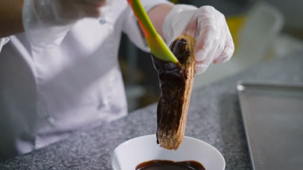 Koka konditorivaror, i en vit kostym och handskar på händerna, smetar den smält choklad gul grön silikon spateln på ett nybakat eclairs. Choklad rinner i en vit skål, som står under éclair — Stockvideo