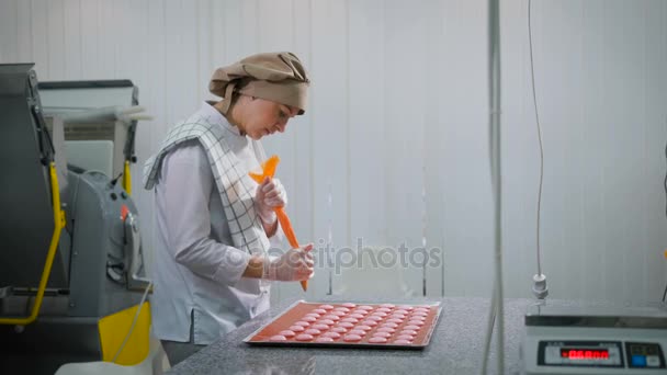 Rodzaju cukiernik młoda kobieta, która stoi bokiem w fabryce wyrobów cukierniczych. Kończy proces formowania połówki różowy makaronik — Wideo stockowe