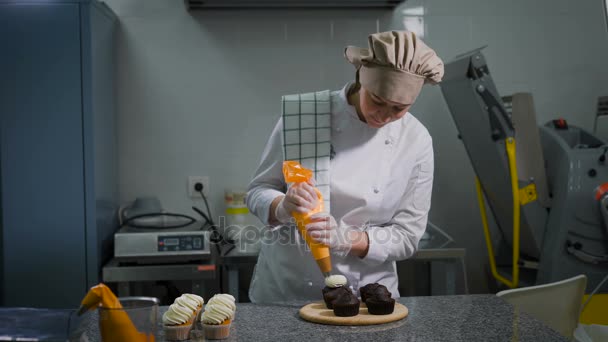 Pastelería femenina exprimiendo suavemente la crema de leche blanca de una bolsa de alimentos para cupcakes de chocolate ya hechos, de pie en un tablero de madera — Vídeos de Stock