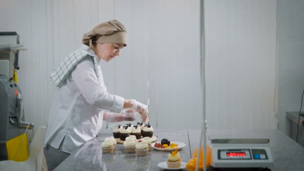 Η γυναίκα μαγειρεύει κέικ για διακοπές. Το κορίτσι η σειρά ως το ψιλικατζίδικο είναι μικρές επιχειρήσεις. Τα κέικ είναι έτοιμο σε ένα τραπέζι. Σε ένα πίνακα cupcakes σοκολάτα και φρούτα. — Αρχείο Βίντεο