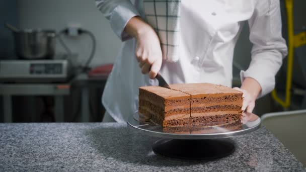 Keuken. Klaar op een tafel chocoladetaart. De werknemer van een snoepwinkel in een witte speciale vorm door middel van een mes snijdt een dessert. — Stockvideo