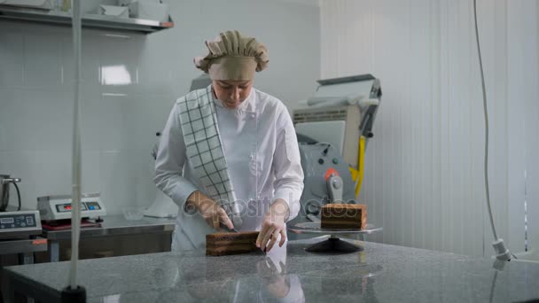 La ragazza in uniforme bianca taglia ordinatamente la torta al cioccolato in tre pezzi uguali in cucina al forno. Panettiere al lavoro guardando di lato . — Video Stock