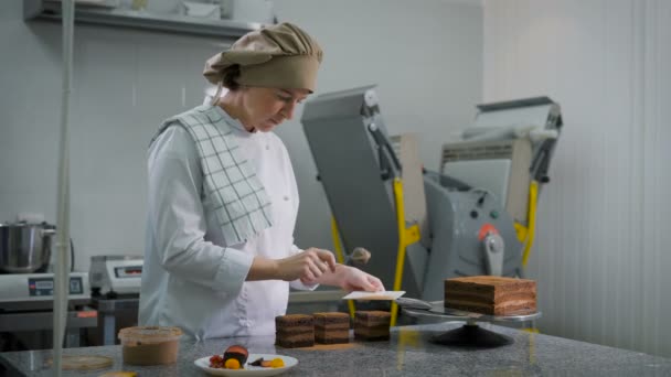 Konditorin in weißem Anzug und brauner Mütze, mit ernstem Blick bestreut, Schokoladenkuchen, die auf dem Tisch stehen, Kakaopulver — Stockvideo