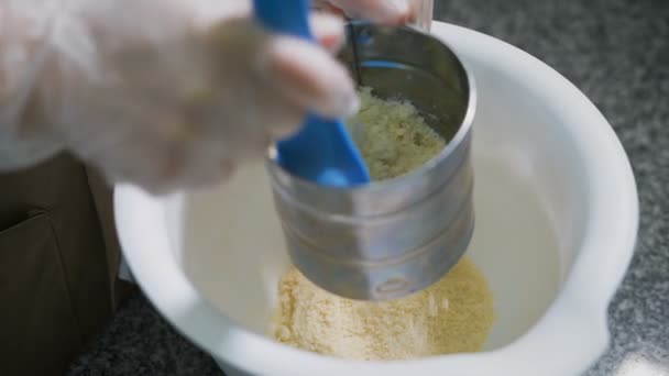 Mestre confeiteiro, cujas mãos em luvas transparentes para a higiene peneira a farinha em uma caneca de metal especial - uma peneira para peneirar a farinha, ajudando-se a uma espátula de silicone azul — Vídeo de Stock