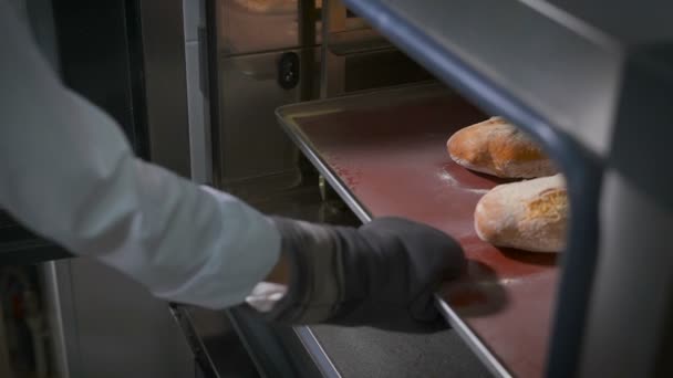 Boulangerie, sur la main de laquelle les gants épais spéciaux, sort de la plaque de cuisson avec du pain fraîchement cuit et une petite coupe au milieu — Video