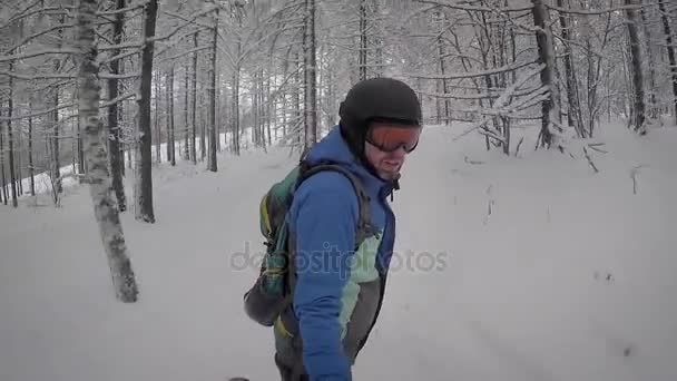 Adult hane med rödaktig skägg som omfattas av rimfrost på en svart snowboard ridning i snörik vinter skog. Bakom honom rider en annan snowboardåkare långt borta, klädd i en ljus jacka och byxor — Stockvideo