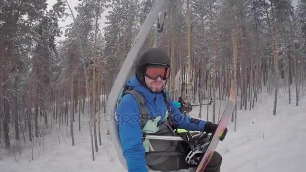 Erwachsener bärtiger Mann, bekleidet mit blauem Snowboardanzug, schwarzem Helm für den Kopf, orangefarbener Sonnenbrille, sitzt auf einem mechanischen Lift, der ihn den Berg hinauf bringt, um dann mit ihr auf einem Snowboard auszuziehen — Stockvideo