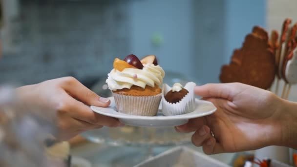 Кондитерша с цветными ногтями посылает маленькую белую руку тарелку, на которой есть два вкусных торта. Один коричневый, другой украшен сливочным кремом и кусочками фруктов — стоковое видео