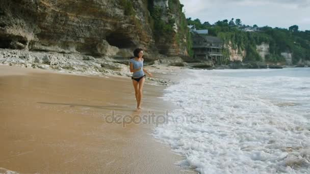 Playa de Bali Island. Sonrisa morena corre a través de la arena en la playa y refrescar los pies en el agua. Mujer disfrutando del sol, océano azul. Con el telón de fondo de impresionantes vistas — Vídeo de stock