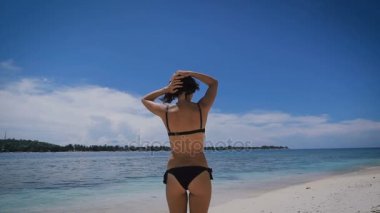 Sırtını kameraya ayakta ve mesafe içine bakarak ince rakam tabaklanmış kız. Kıvırcık Kumral saç kumlu plaja yakın mavi okyanus üzerinde düzeltiyor.