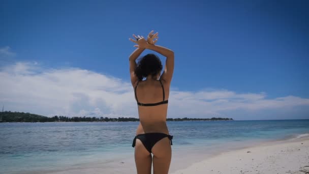 Chica joven y sexy en bikini negro de pie en la playa, levantando las manos y bañándose al sol en la arena de la isla de Bali. Morena en traje de baño negro se broncea y respira aire fresco del mar de Indonesia . — Vídeo de stock