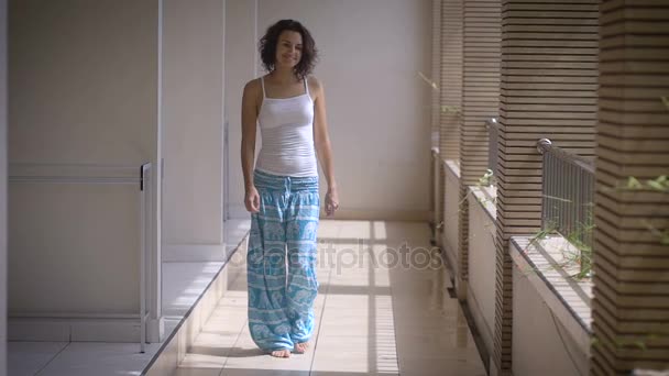 Recreación. Linda chica en el pasillo del hotel. Morena caminando a lo largo del pasillo y camina hacia los postes del balcón. Una mujer con una camiseta y pantalones azules sueltos y su buen humor . — Vídeos de Stock