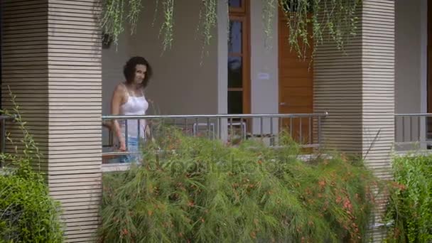 Beautiful brunette walking out on balcony of her tropical private villa and breathing in fresh Balinese air. Young female traveler enjoying her vacation on terrace among tropical luxuriant vegetation. — Stock Video