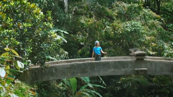 Mujer viajera con ropa azul casual girando en el puente y sosteniendo selfie-stick en sus manos. Atractiva morena haciendo foto panorámica dentro de una exuberante vegetación en la selva tropical . — Vídeo de stock