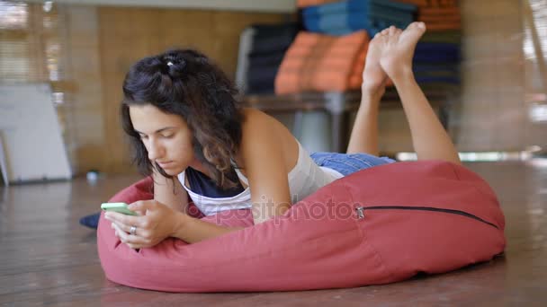 Beautiful girl lying on a large pillow on a tropical resort in Bali. Young woman use smartphone while relaxing in the hotel. Brunettes hair blowing wind. — Stock Video