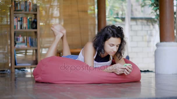 Joven mujer morena acostada en una bolsa de frijoles rosa brillante en un balcón de la casa tropical usando su teléfono inteligente. Hermosa chica relajándose en su casa charlando y haciendo sonar su teléfono en la bolsa de almohada grande . — Vídeos de Stock