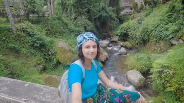 Kadın Gezgin selfie-sopa onun elinde tutan köprü kenarına oturmuş. Çekici esmer arkasından yüzen Nehri ile tropikal orman fotoğrafı panoramik selfie yapmaya çalışıyorum.