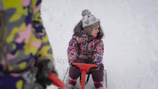 Μητέρα βόλτα ένα μικρό κορίτσι σε ένα έλκηθρο με ένα τιμόνι σε ένα μονοπάτι χιόνι στο πάρκο. Ευτυχισμένο παιδί κοιτάζει γύρω κατά τη διάρκεια μια βόλτα στο φρέσκο αέρα. — Αρχείο Βίντεο
