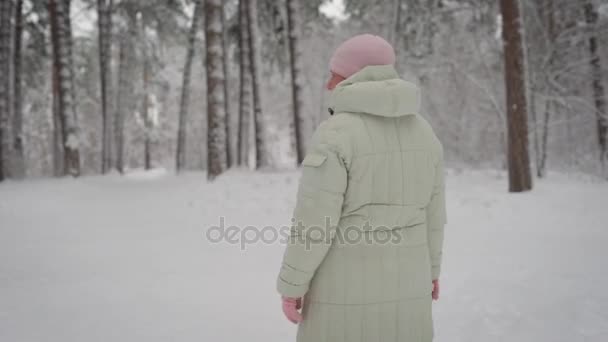 Winter im Wald. Die alte Frau spaziert am Nachmittag auf einem Fußweg. Der Rentner in warmer Jacke und pinkfarbener Mütze verbringt seine Freizeit mit Spaziergängen. atmet frische Luft. — Stockvideo