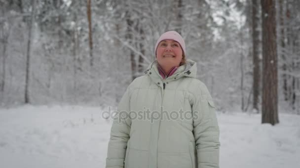 Щаслива жінка в бежевому пальто і рожева шапка, що стоїть в лісі, дивлячись вгору і посміхаючись. У літньому віці жінка-пенсіонер відпочиває в зимовому лісі, поклавши руку їй на голову і задоволений погодою . — стокове відео
