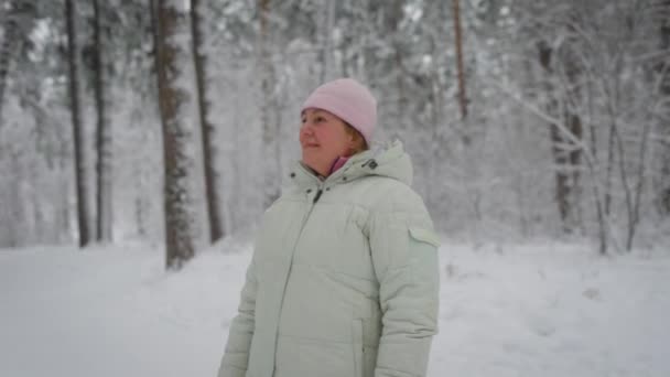 Mujer adulta con cabello rubio, que se esconden bajo el sombrero rosa, con una chaqueta blanca de invierno, de pie en medio de un bosque de pinos cubierto de nieve y disfruta de un hermoso día helado — Vídeos de Stock