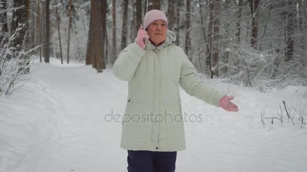 Mujer amable, vestida con un sombrero de color rosa brillante y guantes, chaqueta blanca larga con capucha, pantalones negros, amigo que habla divertido en el teléfono inteligente, diciendo lo que alrededor del paisaje del bosque de invierno, caminos nevados . — Vídeos de Stock