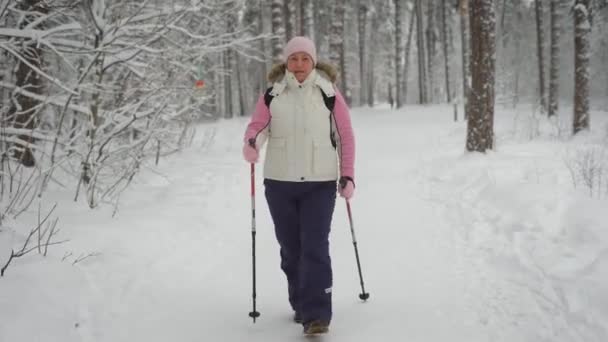 Mujer activa en ropa de abrigo haciendo caminatas nórdicas en el bosque. Mujer deportista feliz pisando con bastones de esquí en la naturaleza de invierno al aire libre. Señora siguiendo el camino demostrando un estilo de vida saludable . — Vídeo de stock