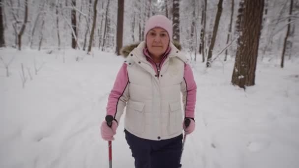 Szép nagymama sétál a havas erdőben, ahol fák, fenyők és más. Ő lendületesen halad előre a görbe mentén. Meleg sportruházat öltözött nő. — Stock videók