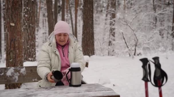 Pensionären i vinter trä. De äldre kvinna sitter på en bänk har resten och kommer att ha varmt te från termos. Efter skandinaviska promenader. — Stockvideo