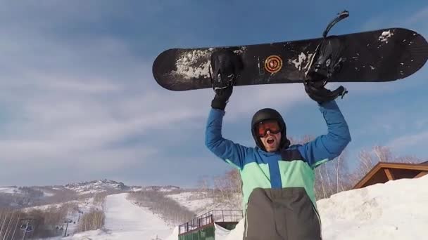 Hombre barbudo adulto de pie en el parque de esquí. Levantó una tabla de snowboard y mostró que estaba contento este día. Él calurosamente vestido con una chaqueta azul, que lo protege del sobrecalentamiento y la humedad . — Vídeo de stock