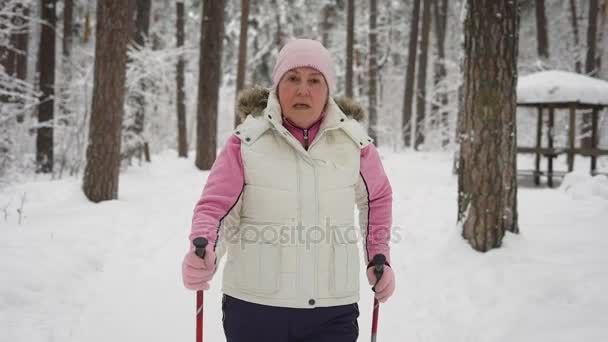 Una donna anziana in un bellissimo abbigliamento sportivo è impegnata nel nordic walking su un percorso innevato nella foresta invernale. pensionato moderno attivamente perseguire il loro tempo libero all'aperto . — Video Stock