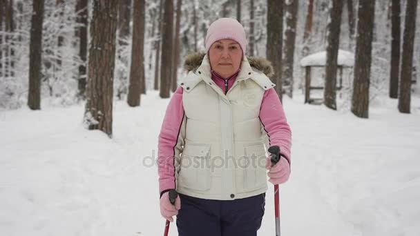 Close-up de mulher ativa gastando seu lazer com exercício ativo caminhada nórdica na floresta. Happy aposentado senhora pisando rapidamente com bastões de esqui no inverno ao ar livre olhando para a câmera . — Vídeo de Stock