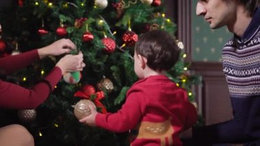 Genç bir aile Noel için evde hazırlanıyor. Uzun saçlı esmer Noel ağacı süsleme. Çocuk oyuncak anne ve asmak için çocuğa yardım etmek çalışan bir kadın atıyor
