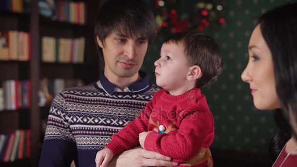 La giovane famiglia a volte guarda da una parte e parla nel soggiorno della sua accogliente casa. La gente magnificamente vestita in previsione delle vacanze di anno nuovo passa il tempo insieme . — Video Stock