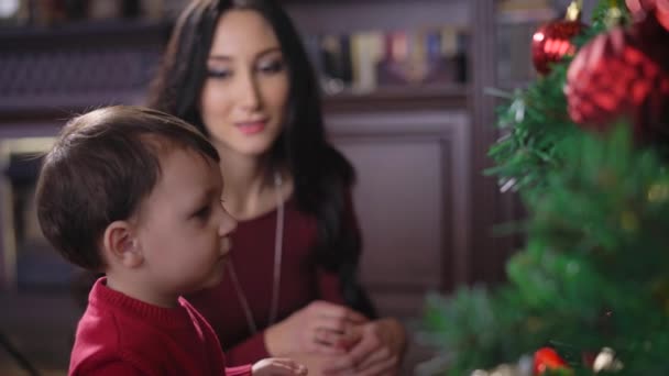 Una chica con un niño en casa en el árbol de Navidad. Morena mirando al niño y sonriendo. El chico juega con un árbol de Navidad. La Navidad es una de las fiestas más cálidas, familiares y espirituales . — Vídeo de stock