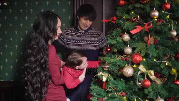 彼の幼い息子に美しく服を着て母親と父親を異なるおもちゃモミの木で飾られています。クリスマスイブに家で若い家族の共同娯楽. — ストック動画