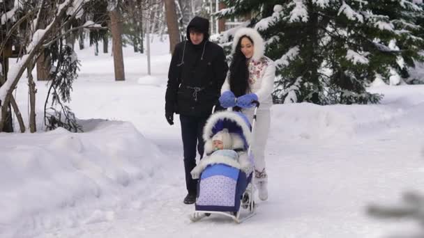 若く美しく着てスキーで、ベビーカーに座ってあなたの子供と雪に覆われた公園の上を歩いての両親。ブルネットは彼女の髪を整えたりと笑顔. — ストック動画