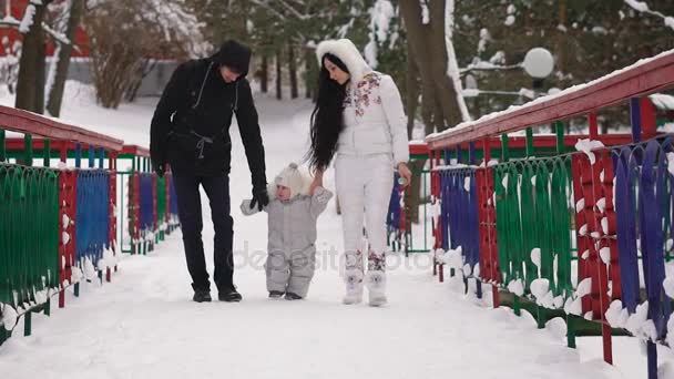 Hombre y mujer sostienen las manijas de su hijo pequeño, que solo recientemente había aprendido a caminar, por lo que lo hace torpemente. Los padres están observando cuidadosamente a su hijo, porque no está tropezado. Familia caminando en el parque de invierno — Vídeo de stock