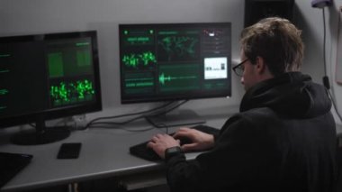 Yeraltı örgütü. Hacker kritik bilgileri kırmaya çalışan. İki bilgisayar ve laptop Masası üzerinde. Bilgi ve bir lamba ile duvar gemide. Adam cidden üzerindeki çalışmasını ayarlanır.
