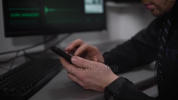 Κοντινό πλάνο της Ρωσικής χάκερ κάθεται στο τραπέζι και τη χρήση του smartphone. Αρσενικό κατάσκοπος hacking σύστημα υπολογιστή με τη βοήθεια του κινητού και λήψη σημαντικών δεδομένων αρχείου από το δίκτυο. — Αρχείο Βίντεο