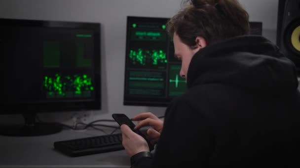 Hombre adulto Hacker, con barba en la cara, con una chaqueta deportiva negro se sienta en un búnker secreto y hace ataques cibernéticos. En su teléfono inteligente, dijo que los datos — Vídeos de Stock