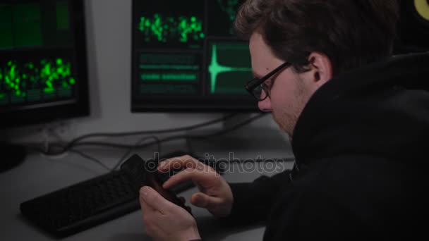 The underground organization. Two computers and a laptop on desk. A man in a dark jacket and glasses looks at the article on your smartphone and tries to understand the information — Stock Video