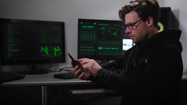 Un hacker en la sala de computadoras. Un hombre en gafas de sol y con un teléfono inteligente en la mano tratando de hackear la información de bases extranjeras. La habitación está equipada con la mayoría de los ordenadores . — Vídeo de stock