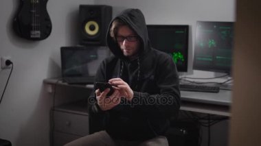 Bir siyah ceket ve kaputun üstünde onun baş, gri pantolon var olduğu birden fazla monitör, dizüstü bilgisayar, müzik için hoparlörler stolon, bir sandalye üzerinde oturan bir adam. Duvarda asılı bir gitar