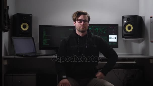 Серьезный хакер смотрит прямо на снимок, он одет в темную куртку, очки, руку, умные часы, каштановые волосы. На заднем плане находятся экраны компьютера, ноутбука, музыкальные спикеры для голоса — стоковое видео