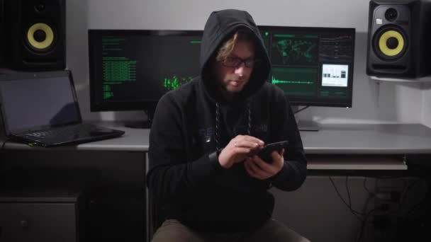 Un hombre con una chaqueta y una capucha en la sala de ordenadores. Un hacker en gafas de sol y con un smartphone a mano sentado en una silla. El tipo escanea nerviosamente e ingresa la información en el teléfono inteligente . — Vídeo de stock