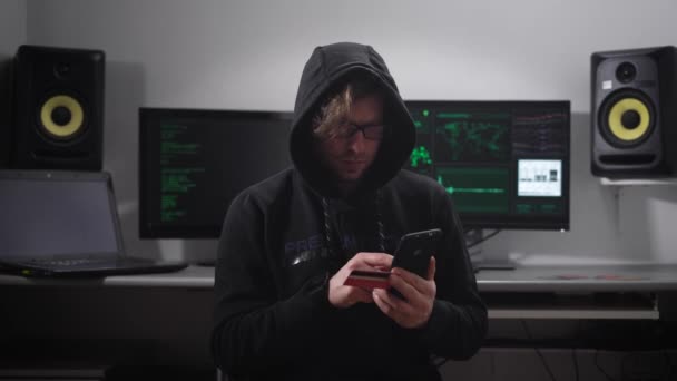 Опытный хакер взламывает банковский счет клиентов, используя смартфон и пластиковую кредитную карту. Молодой человек в куртке с капюшоном занимается незаконной деятельностью в сфере ИТ . — стоковое видео