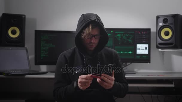 Primer plano de un hacker encapuchado haciendo ciberataque en la red bancaria con la ayuda de un teléfono inteligente en sus manos. Hombre de negro está escribiendo datos de robo rápido de tarjetas de crédito . — Vídeos de Stock